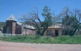 Image of a small, worn, brick church with a white picket fence surrounding on the left. to the right is a yellow house with overgrown weeds in front. A hill is behind the two buildlings and a blue sky above.