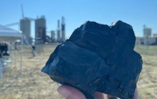 up close image of a lump of coal in a person's hand with a power plant in the background on a sunny day