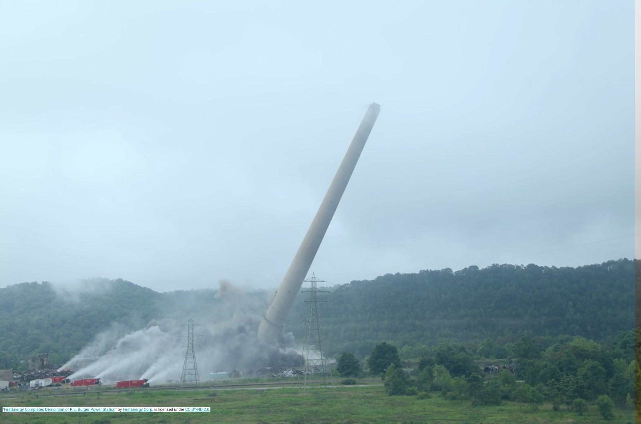 Coal powerplant tower collapsing in a demolition, surrounded by spraying fire engines. Background is a green hill and cloudy sky.